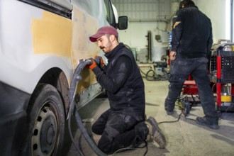 Male auto body worker kneeling and using a power sander with vacuum attachment on a van door,