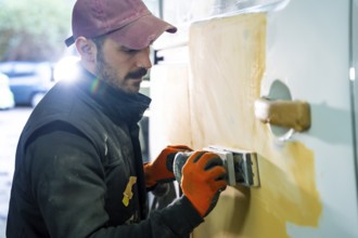 Skilled auto body worker wearing work gloves and a cap carefully sanding a car door with filler
