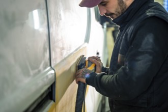 Mechanic working carefully, sanding a vehicle body panel with an orbital sander to prepare it for
