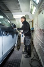 Auto body technician cleaning and preparing a dark colored car for painting inside a professional