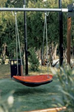A bright orange hanging swing is set in a serene garden surrounded by trees and grass. The peaceful