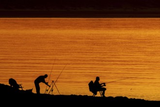 Silhouette of two recreational anglers, fishermen with several fishing rods angling at sunset