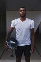 A focused American football player stands outdoors holding his helmet, wearing a white jersey. His