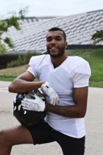 A confident American football player in uniform poses outdoors, holding his helmet. The setting