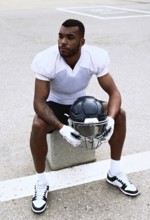 An american football player in a white jersey sits on a bench holding his helmet, reflecting before