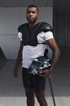 A focused football player stands with his helmet, dressed in shoulder pads and uniform,