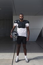 A determined American football player stands confidently, wearing shoulder pads and holding a
