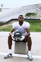 American football player sits on a stone block, holding a helmet. The scene is set in an urban park
