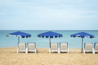 A serene beach scene with three blue umbrellas and white lounge chairs on golden sand facing a
