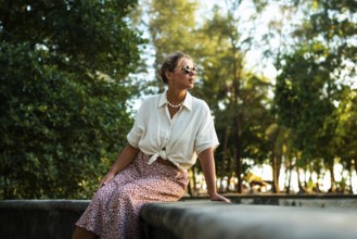 A stylish woman in sunglasses and summer attire sits on a stone ledge surrounded by lush greenery