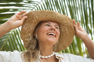 A joyful woman wearing a straw hat and shell necklace smiles under palm leaves, capturing a perfect
