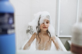 A joyful child is playing with bubbles in a bathtub, with a big smile and wet hair. The scene