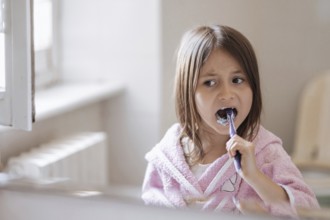 A young girl wearing a pink bathrobe brushes her teeth with a toothbrush, highlighting morning