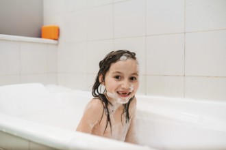 A cheerful child bathes in a white tub, surrounded by soap bubbles and foam. The joy of bath time