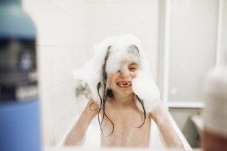 A joyful child enjoys a playful bath, covered in foamy soap bubbles, smiling widely. The bright
