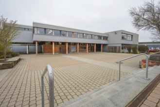 A schoolyard with extensive pavement, gray building structure and outdoor tree, Schlehengäu Grund