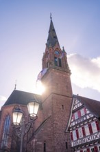 Church tower in sunlight next to a traditional half-timbered house under a clear sky, Calw, Calw