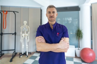 Confident male physiotherapist or osteopath standing with arms crossed in clinic, providing