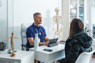 Male doctor in scrubs and stethoscope is talking with a female patient seated across his desk,