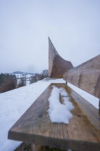 Wooden bench in snow with modern building in background and winter atmosphere, Sinnraum, Oberreute,