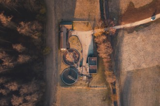 Sewage treatment plant with round basin and buildings from above, surrounded by fields and forest