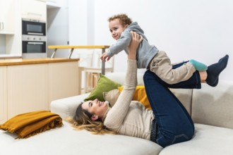 Mother lying on a sofa at home, joyfully playing with her young son, lifting the toddler up in the