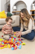 Mother and young son are sitting on the floor at home, building a tower with vibrant wooden blocks,