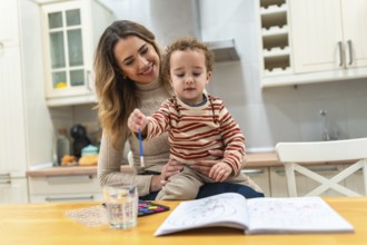 Mother holding her young toddler on her lap, together they are painting in a coloring book with