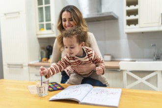 Happy mother holding son at the kitchen table as he dips a paintbrush into water, preparing to