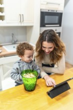 Mother and young son enjoying a family moment, sharing popcorn while engaging with a digital tablet