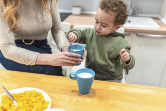 Mother guiding her toddler son stirring milk in a cup during breakfast, creating a moment around