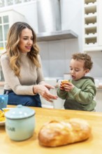 Mother looking at her happy child holding a muffin while having breakfast in a home kitchen,
