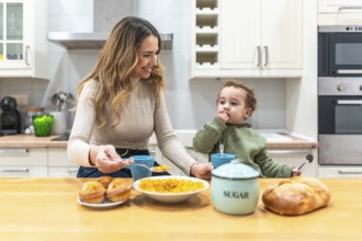 Young smiling mother feeding cereal to her adorable toddler son while sitting at the kitchen table,