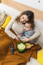 Mother holding her young son while they are relaxing on a sofa under a cozy blanket, watching a