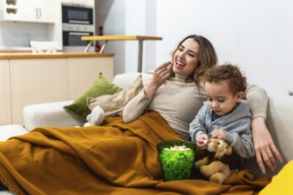 Mother and young son relaxing on a sofa under a blanket, eating popcorn and watching a movie,