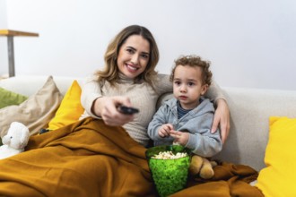 Mother and child relaxing on a cozy sofa covered by a blanket, enjoying popcorn and bonding