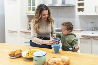 Mother helps young son eat cereal at a wooden kitchen table in a sunlit morning, sharing a cozy,