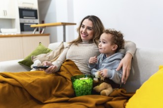 Mother and young child cozy on the sofa sharing popcorn and a remote, smiling and watching tv
