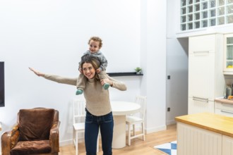Happy mother gives toddler son a piggyback airplane ride in a bright modern apartment, sharing