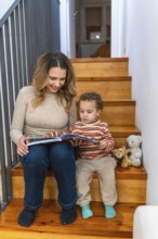 Happy mother and toddler boy enjoying reading a colorful storybook together while sitting on wooden