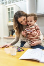 Mother holding her toddler son while they are painting with watercolors at a wooden table in a