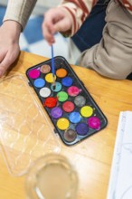 Child engaging in creative activity, dipping a small paintbrush into a colorful watercolor paint