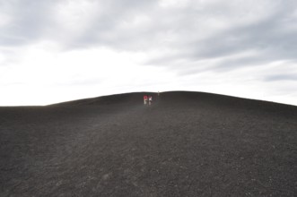 Wide landscape with people walking up a lava hill under cloudy sky, Craters of the Moon National