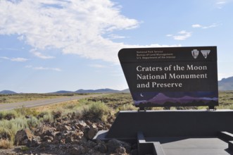 Craters of the Moon National Monument sign next to a road with wide landscape and blue sky, Craters