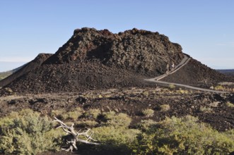 Hikers climb a volcanic mound of lava rock, sparse vegetation along the way, Craters of the Moon