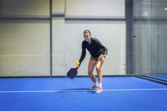 Woman athlete smiling, holding a paddle and yellow ball on a vibrant blue pickleball court,