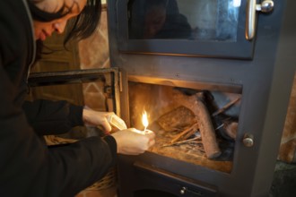 An Asian woman is seen lighting a wood burning stove, carefully holding a match to the wood. She is