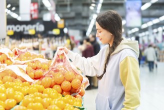 A woman is seen selecting oranges from a fresh produce section in a bustling supermarket, examining