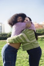 Two friends embracing joyfully in a sunny park, wearing cozy sweaters and jeans. Their happiness is