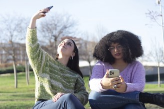 Two friends in the park, one takes a selfie while the other looks at her phone. They are enjoying a